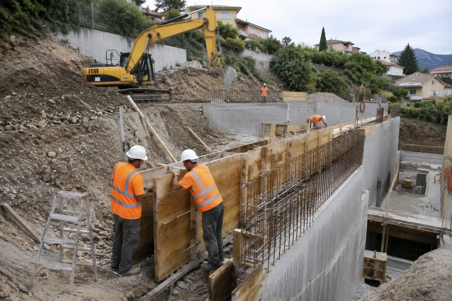 Coffrage béton armé pour un mur de soutènement sur terrain en pente, pelle mécanique en fond de chantier